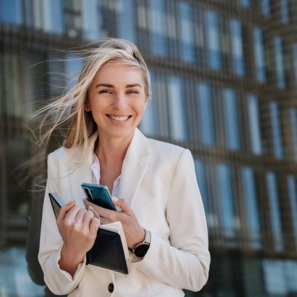 Gorgeous caucasian young woman in white suit with fluttering hair smiling wide, holding diary and phone. Successful manager received great news, financial profit, business growing. Happy businesswoman